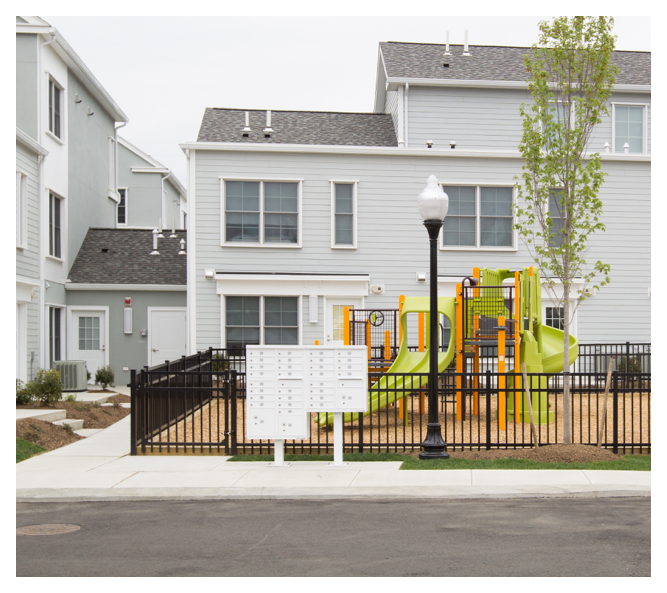 Secure mailboxes sit in front of a gated playground, featuring a colorful playscape on top of a soft bed of wood chips.