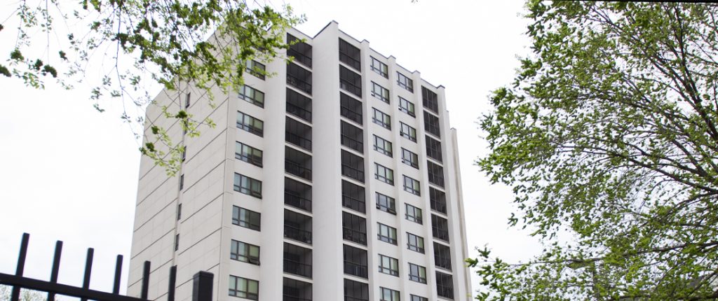 A view peeking through trees, looking up at nine stories of Clinton Manor from the parking lot.