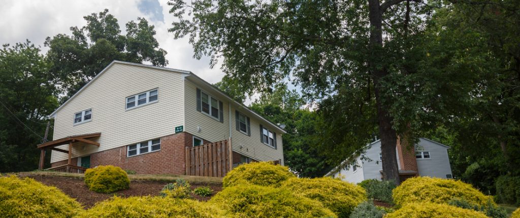 View through landscaped shrubbery up at a two-floor housing unit featuring an attractive exterior of brick and siding.