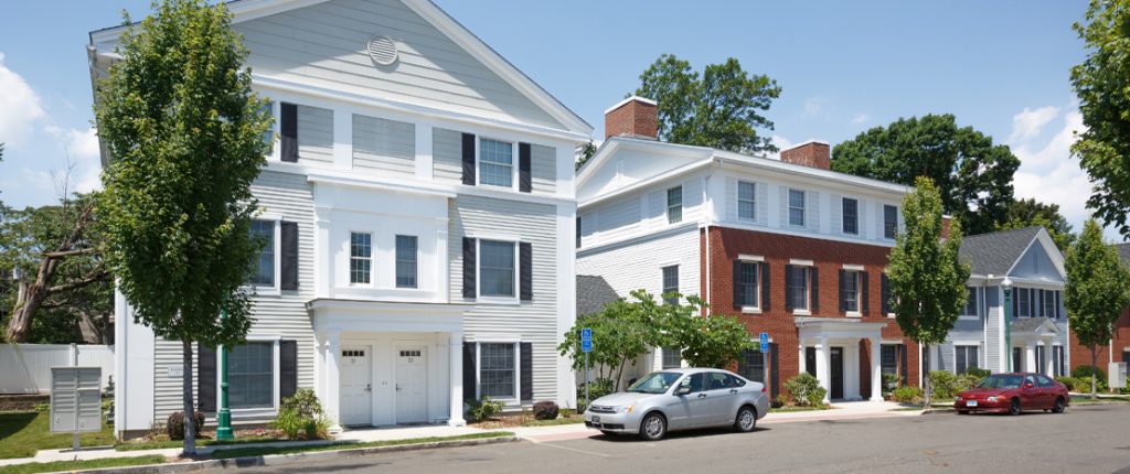 Several Colonial-style buildings sit along a sunny street dotted with trees and historic-looking street lamps.