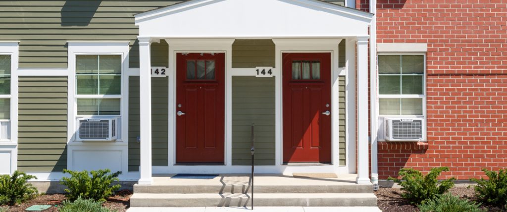 A landscaped covered entrance features Colonial elements and red doors. Window AC units are visible.