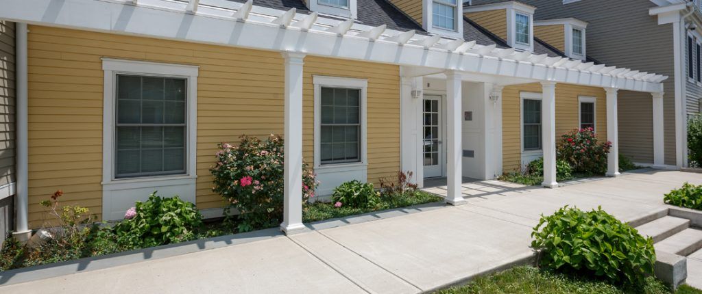 A slat canopy provides partial shade to the plants and flowering bushes lining the side of a pristine building.