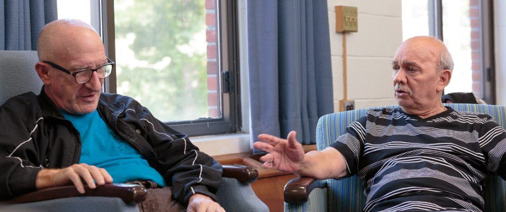 Two senior residents engage in conversion seated in comfortable-looking chairs near a bright window.