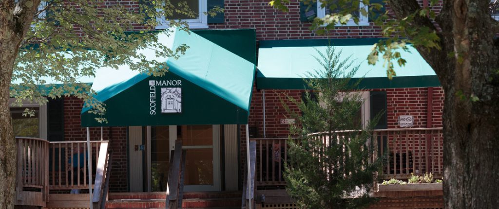 Looking through the trees at the porch-like entrance to Scofield Manor. Residents sit outside shaded by the large awning.