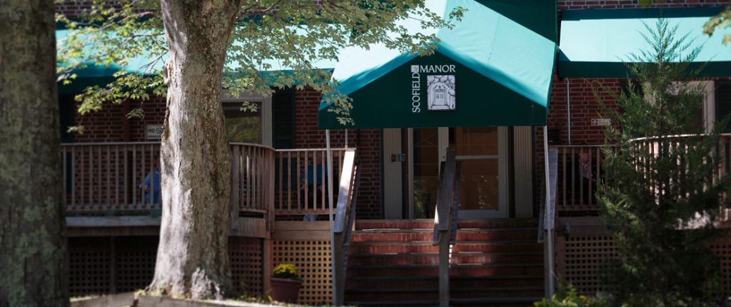 The Scofield Manor porch is lined with wood railings. A large awning provides shade for residents sitting outdoors.