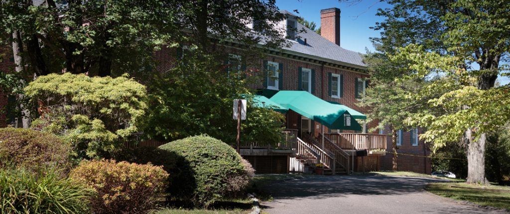 The large brick building, surrounded by trees and shrubbery, looks homey with its many windows and faux shutters.