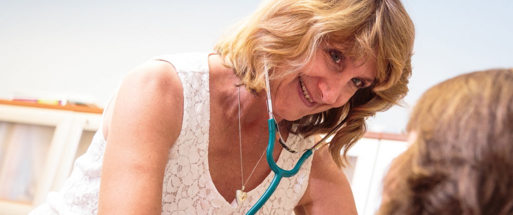 A smiling, on-site registered nurse checks on a resident using a stethoscope.