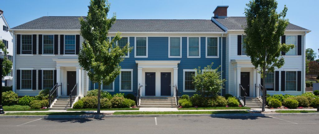 This Colonial-style building is four housing units with large windows and private entrances surrounded by green landscaping.