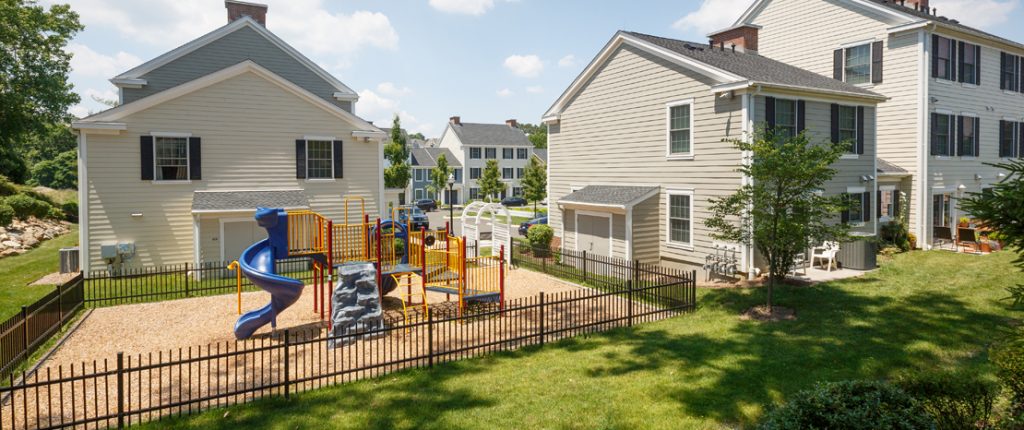 The communal playground features a colorful playscape on a safe substrate, surrounded by manicured grass and small trees.