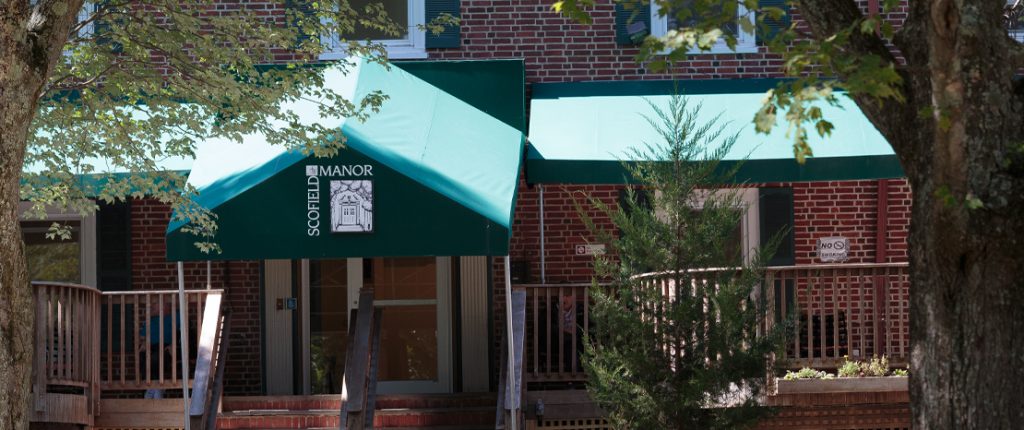 Looking through the trees at the porch-like entrance to Scofield Manor. Residents sit outside shaded by the large awning.