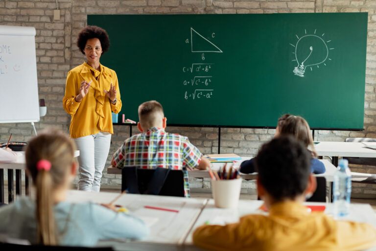 Teacher in a yellow blouse standing in front of a chalkboard, explaining a math lesson to a classroom of students.