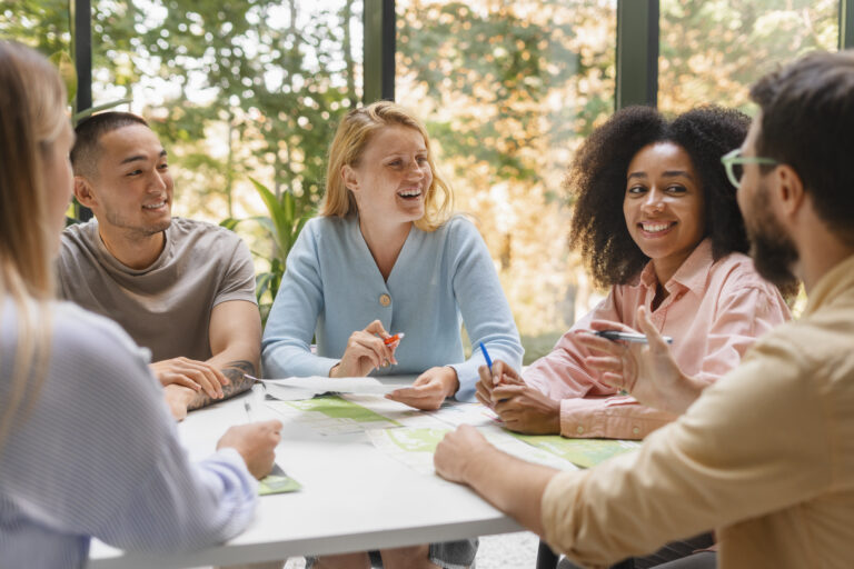 Group of young adults sitting at a table, smiling and collaborating during a discussion in a bright space with greenery outside the windows.