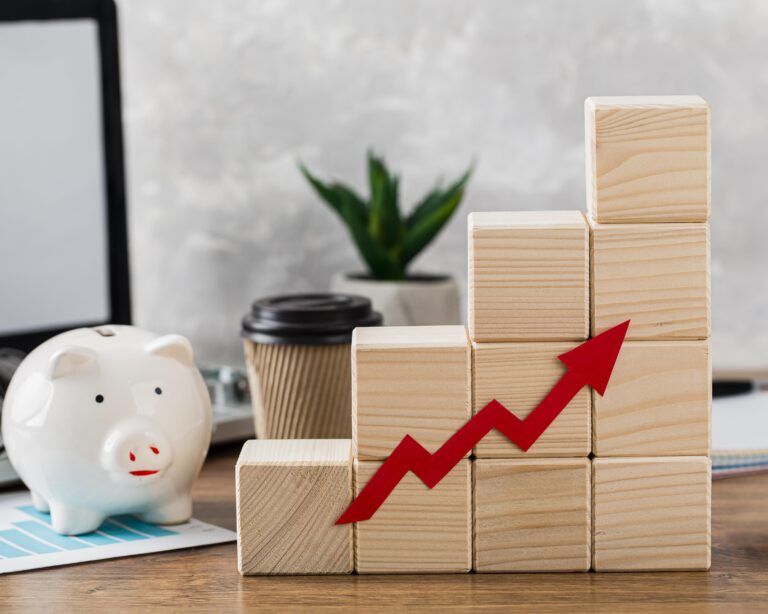 A white piggy bank beside stacked wooden blocks arranged in an upward staircase with a red arrow pointing up