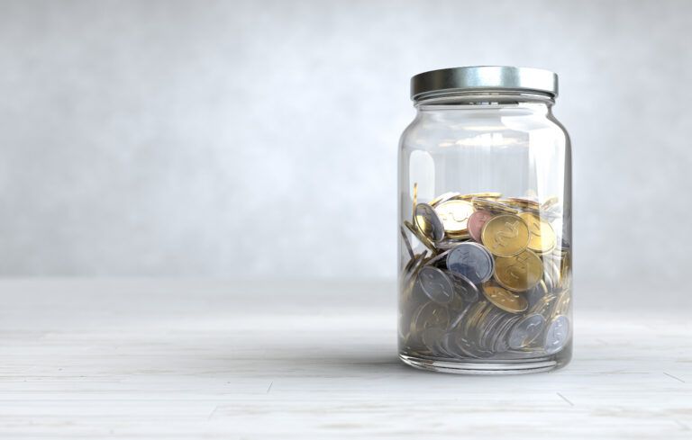 Clear glass jar partially filled with mixed coins sitting on a light-colored tabletop against a neutral background.