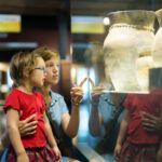 Adult and child standing in a museum gallery, looking at ceramic vessels displayed behind glass.