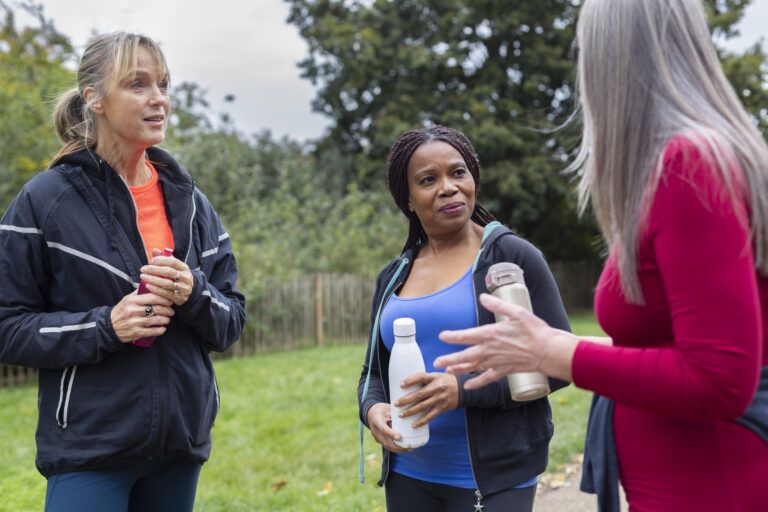 Three women standing outdoors in a park, talking after a walk and holding reusable water bottles.