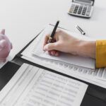 Person writing notes in a planner beside financial documents and a pink piggy bank on a desk.