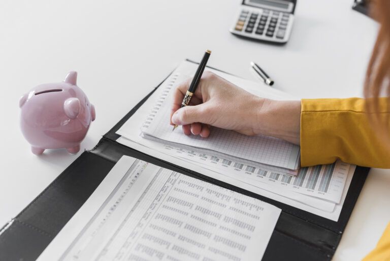 Person writing notes in a planner beside financial documents and a pink piggy bank on a desk.