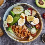 Healthy meal bowl with grilled chicken, vegetables, avocado, eggs, and lemon slices on a table