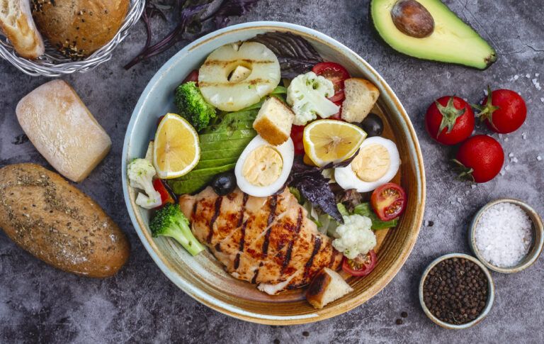Healthy meal bowl with grilled chicken, vegetables, avocado, eggs, and lemon slices on a table